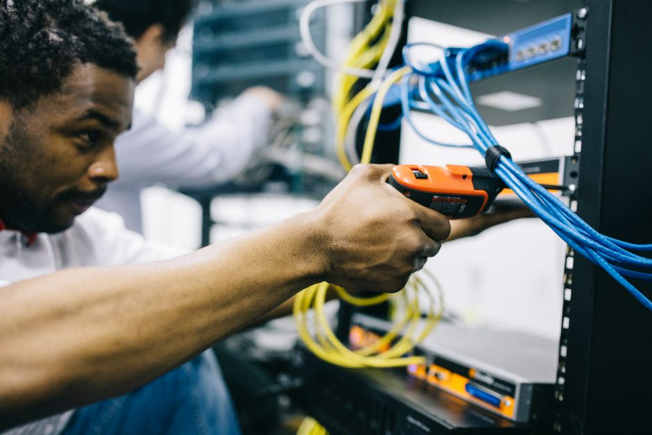 Ingeniero afroamericano concentrado reparando equipo con un destornillador eléctrico, visto de lado